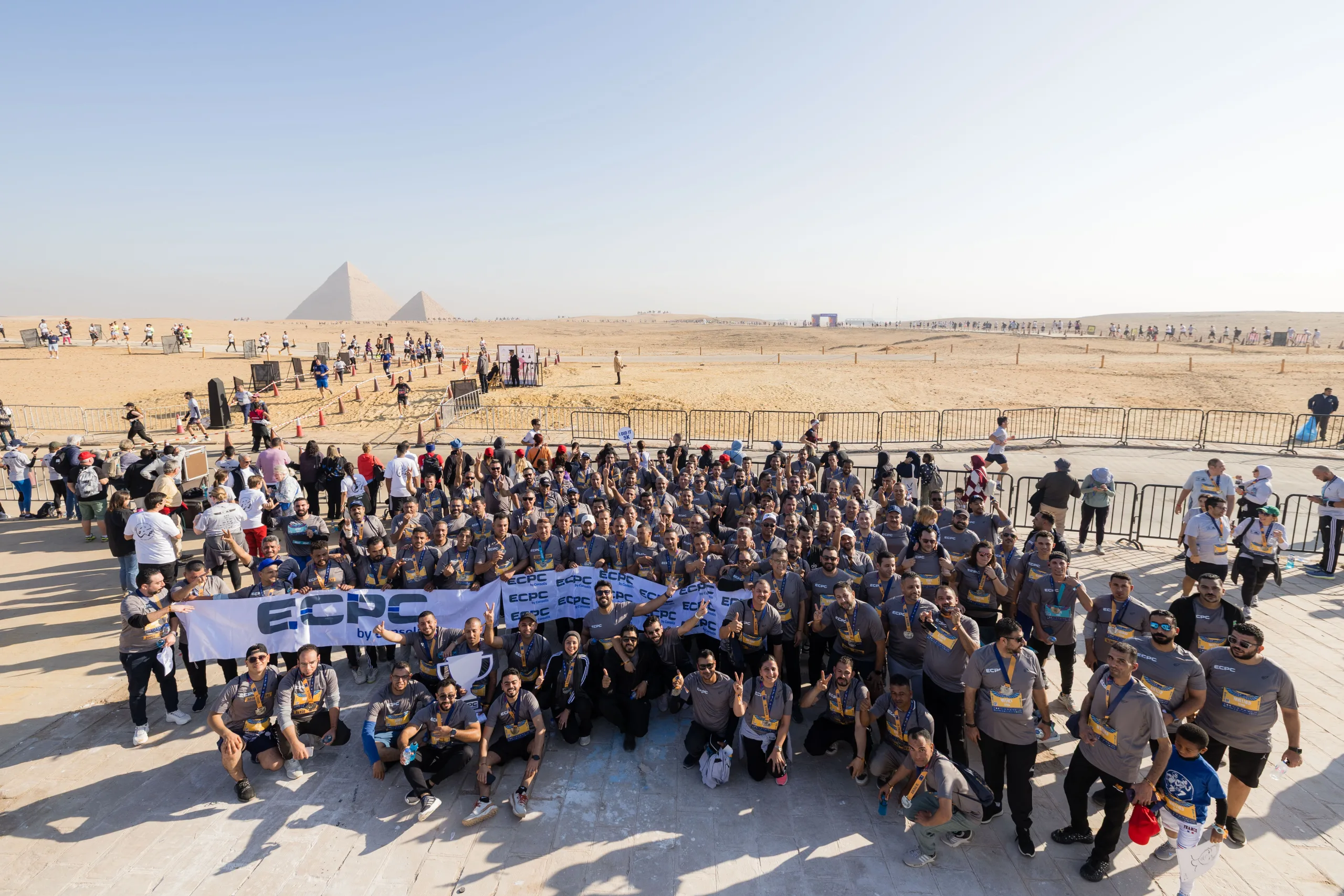 A wide-angle view of a large crowd of ECPC employees in matching grey jerseys holding a banner and cheering at the Giza Pyramids event.