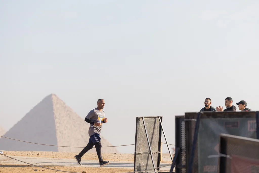 A side profile of a man in a grey ECPC jersey running on a paved track with a massive ancient pyramid visible in the distance.