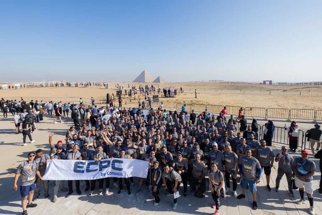 A large group of ECPC employees in grey jerseys holding a banner and trophy in front of the Great Pyramids during the 2025 half marathon.