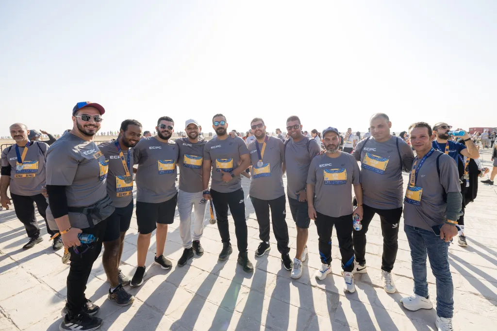 A group of men wearing grey ECPC jerseys and finisher medals standing together on a sunlit paved area at the Pyramids Half Marathon.