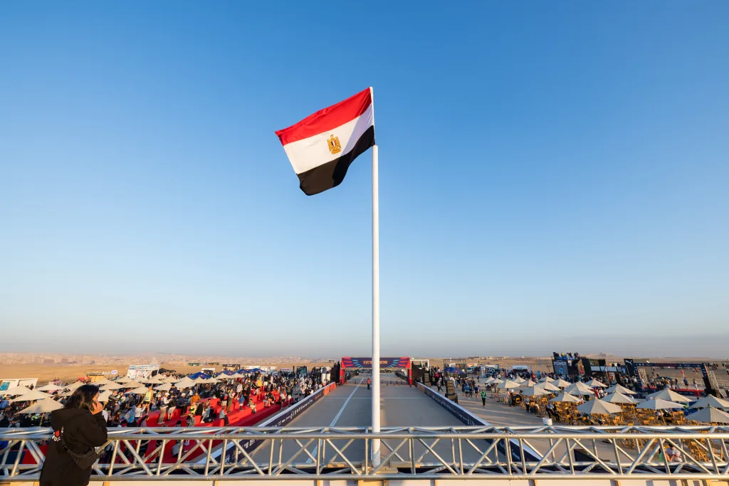A tall flagpole with the Egyptian flag waving in the wind, overlooking the marathon event infrastructure and crowds in the desert.