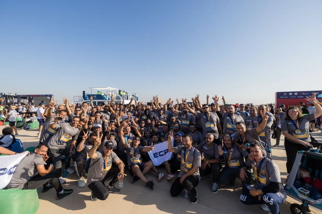 A large group of runners in grey ECPC shirts sitting and standing together, celebrating with finisher medals at the Giza race village.
