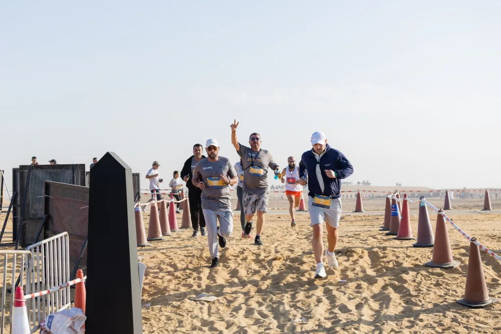 Three men in ECPC athletic gear running on a sandy path lined with traffic cones during a marathon, with one runner waving a peace sign.